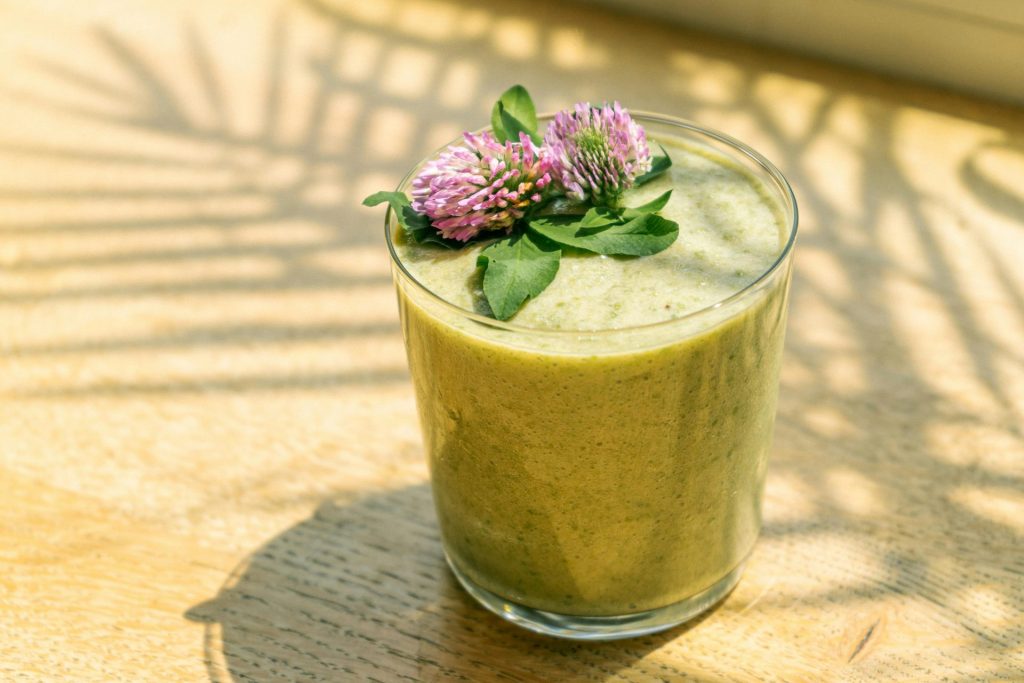 Close-up of a nutritious green smoothie topped with fresh purple clover flowers in sunlight.
